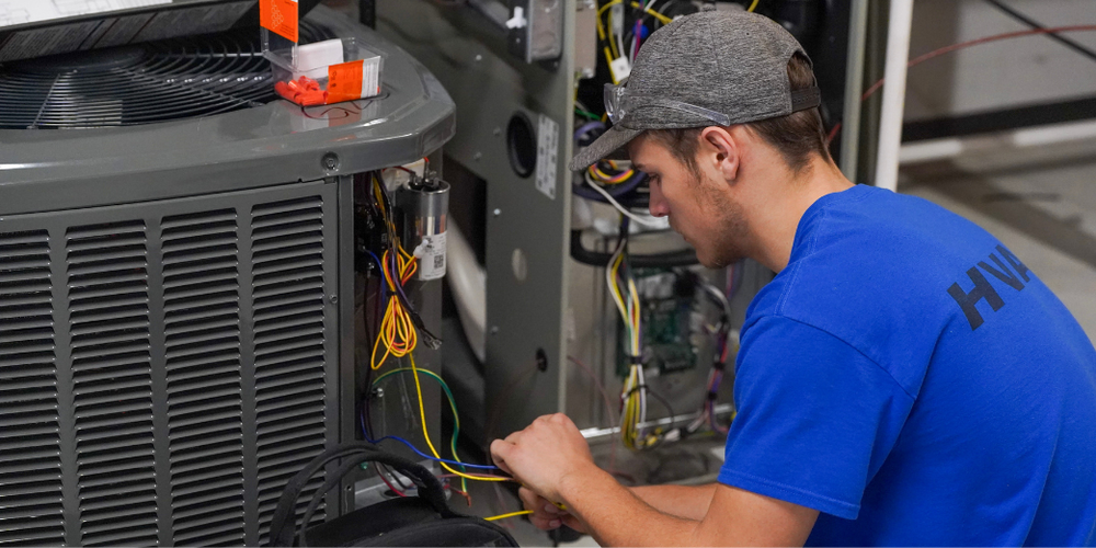 A student in a blue shirt and gray hat works on the wiring of an AC unit.