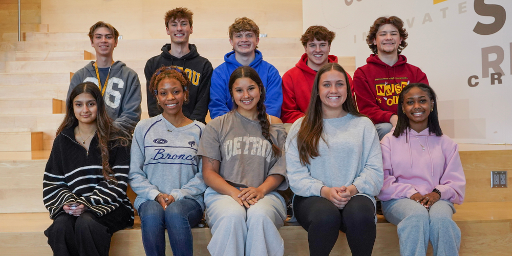 A group of 11 diverse teenagers sitting on bleachers in a gymnasium-like setting. Some wear hoodies and casual clothing. A chalkboard or whiteboard behind them has text visible.