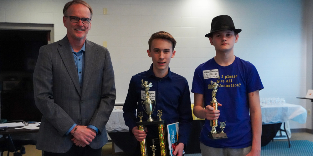A man and two boys stand together smiling and the two boys are holding up trophies