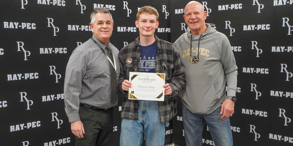 Three men standing together with the man in the middle holding a certificate