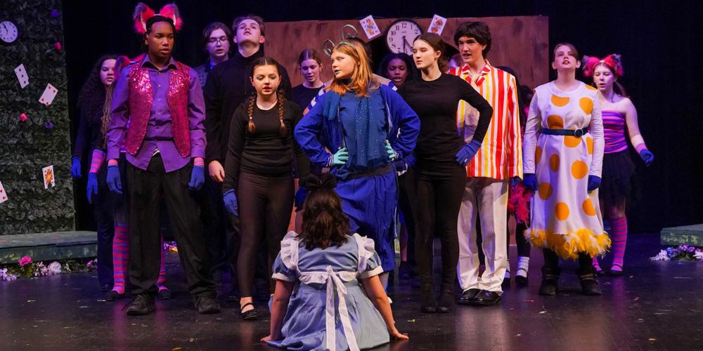 A cast in colorful costumes on stage with a green background.