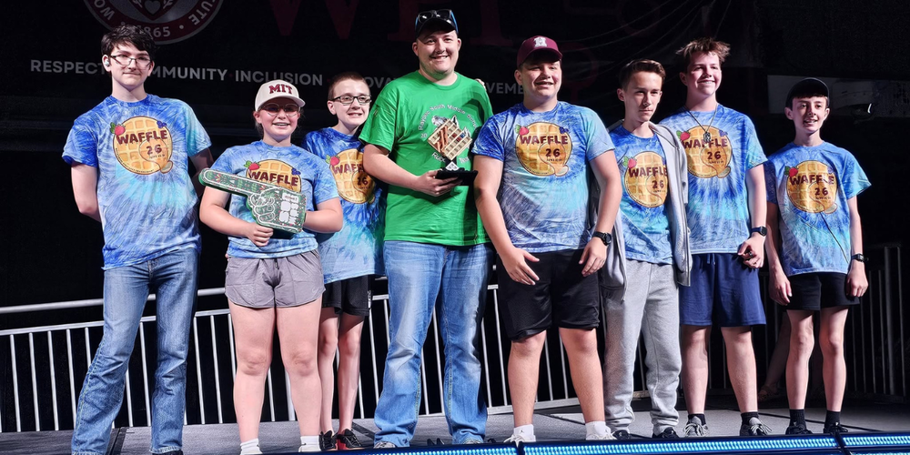 A group of students in blue shirts and a man in a green shirt stand on stage together smiling.