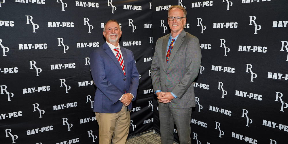 Two men in suits standing in front of a black back drop that says "RP Ray-Pec"