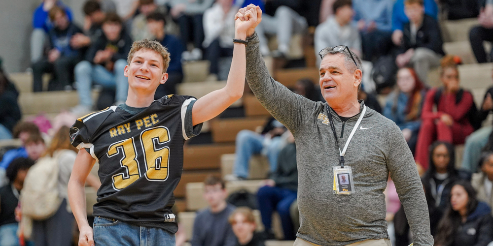 A student in a black and gold jersey with the number 36 smiles as a man holds up his arm in a victory pose