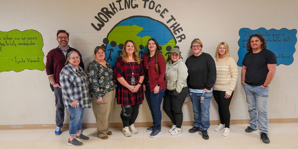 A group of adults stand in front of a brick wall with blue and green paintings as they all smile.