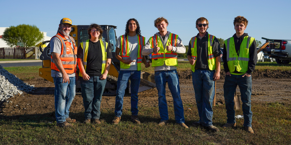 Six construction workers wearing safety vests and hard hats standing in a line on a dirt construction site with equipment in the background.