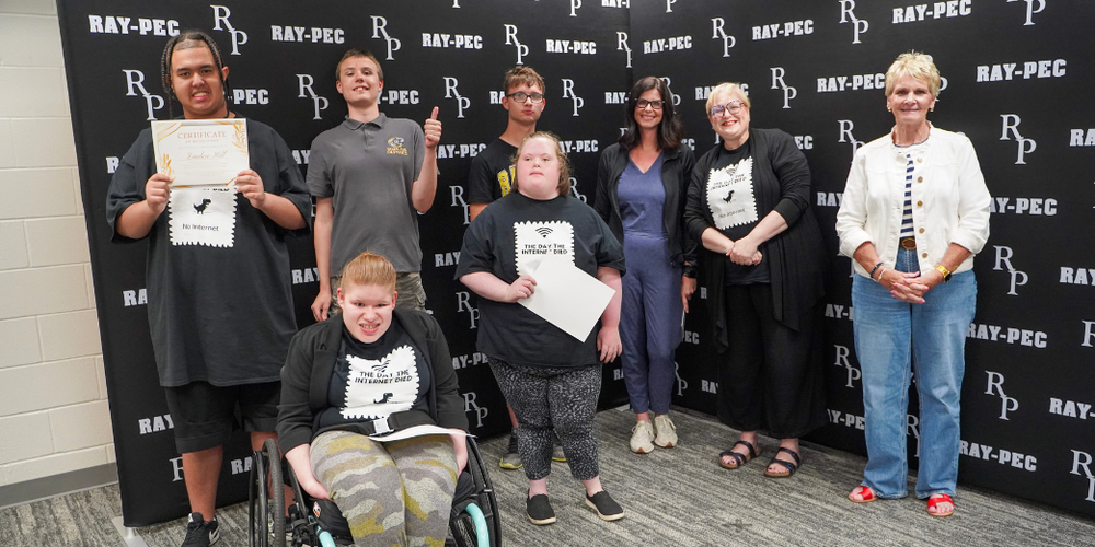 A group of students stand together with three adults holding up certificates in front of an RP backdrop