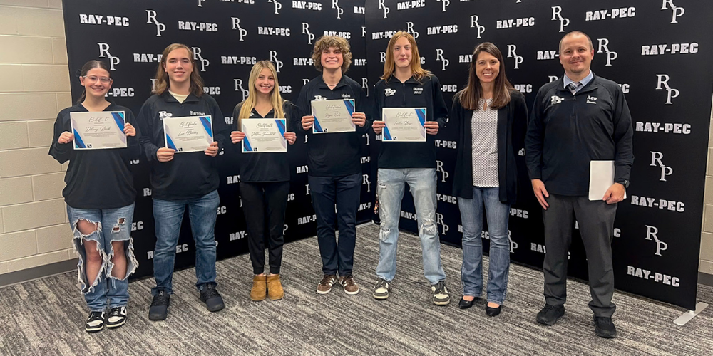 A group of students and two teachers stand in front of a backdrop holding certificates