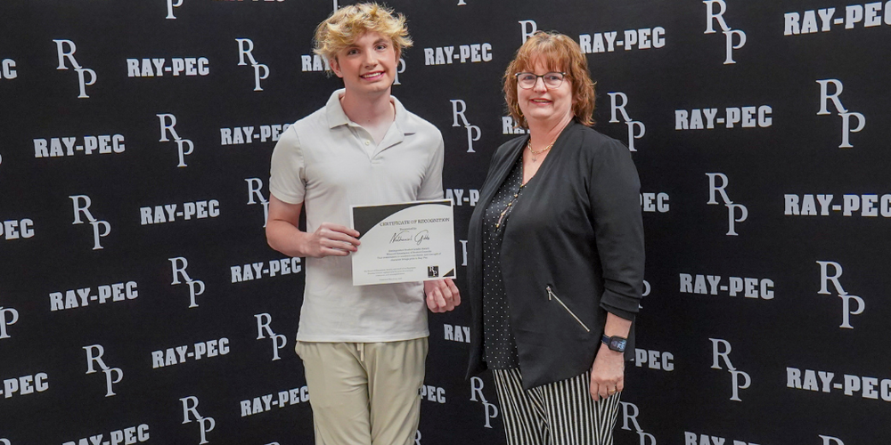 A boy and a woman stand in front of a black backdrop that says, "Ray-Pec" while the boy holds a certificate
