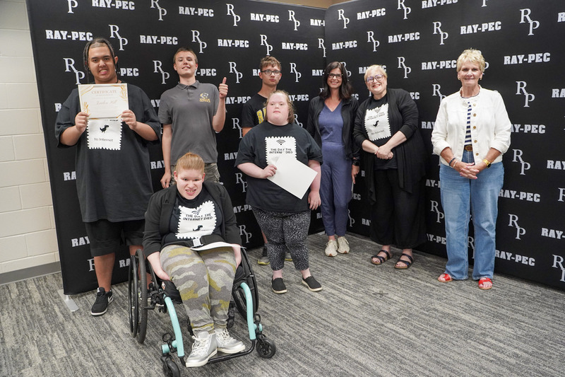 A group of student smile with certificates with three adults, all standing in front of a black RP branded backdrop.