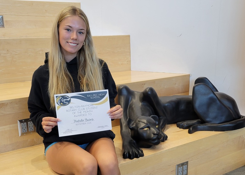 A student holding a certificate, sitting on a stair case with a panther statue next to them.