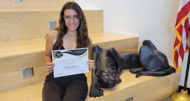 A student holding a certificate, sitting on a stair case with a panther statue next to them.
