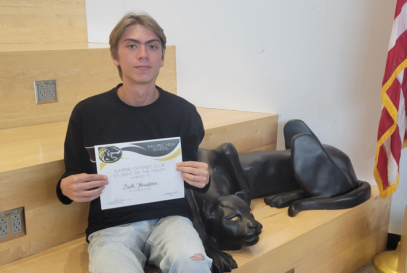 A student holding a certificate, sitting on a stair case with a panther statue next to them.