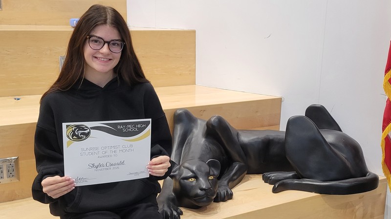 A student holding a certificate, sitting on a stair case with a panther statue next to them.