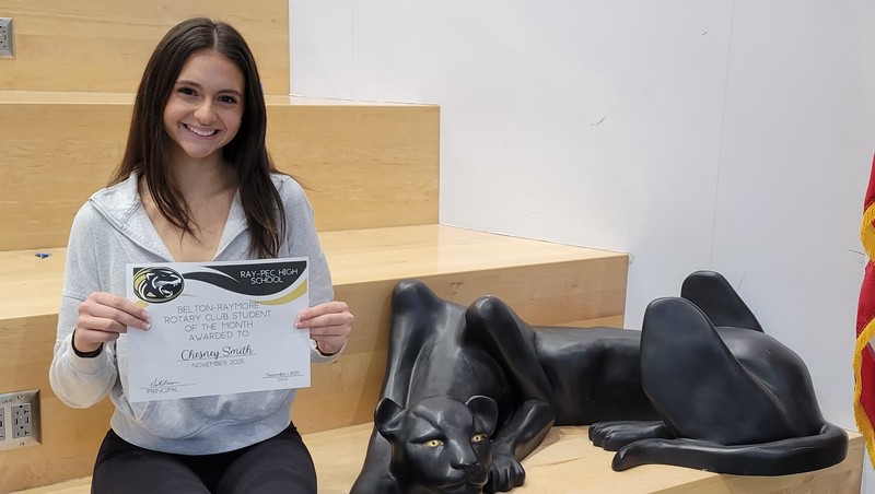 A student holding a certificate, sitting on a stair case with a panther statue next to them.