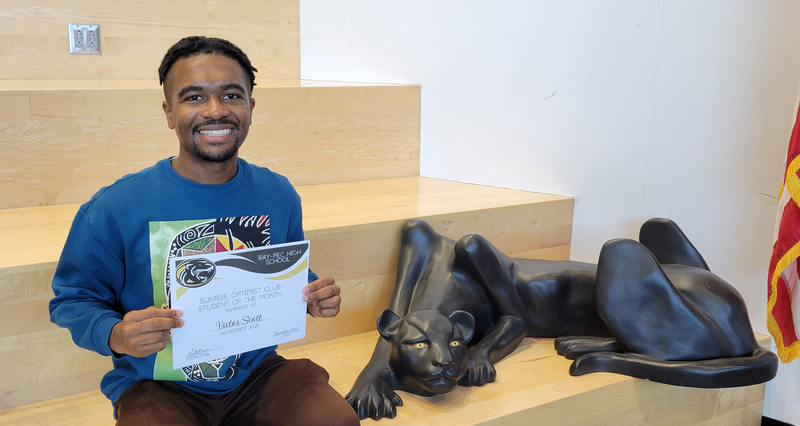A student holding a certificate, sitting on a stair case with a panther statue next to them.
