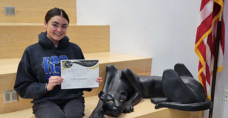 A student holding a certificate, sitting on a stair case with a panther statue next to them.