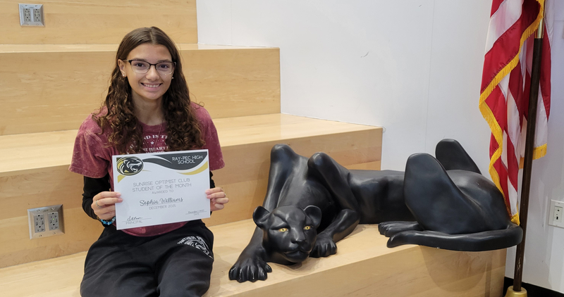 A student holding a certificate, sitting on a stair case with a panther statue next to them.