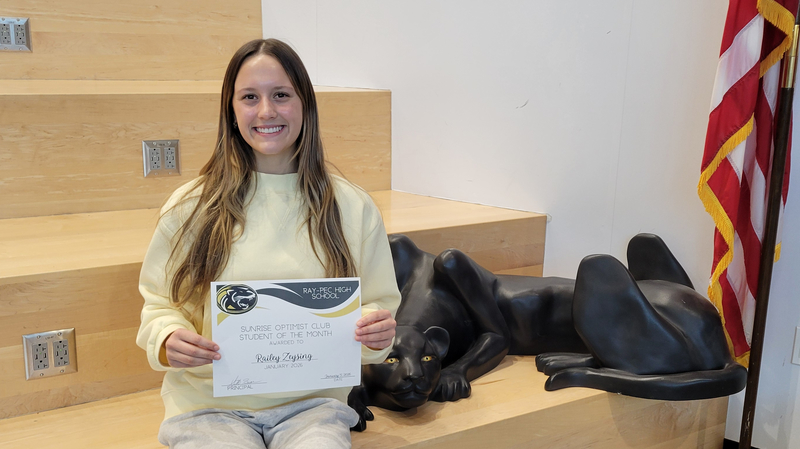 A student holding a certificate, sitting on a stair case with a panther statue next to them.
