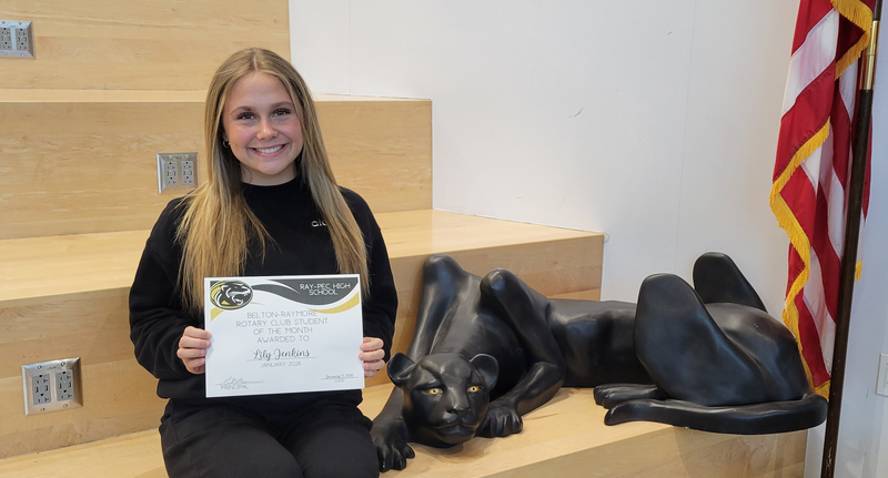 A student holding a certificate, sitting on a stair case with a panther statue next to them.