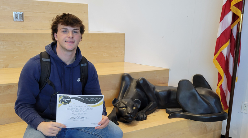 A student holding a certificate, sitting on a stair case with a panther statue next to them.