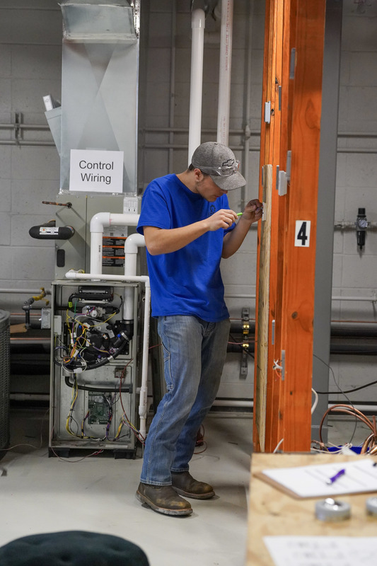 A student in a blue shirt stands and screws in some wires to the wall.