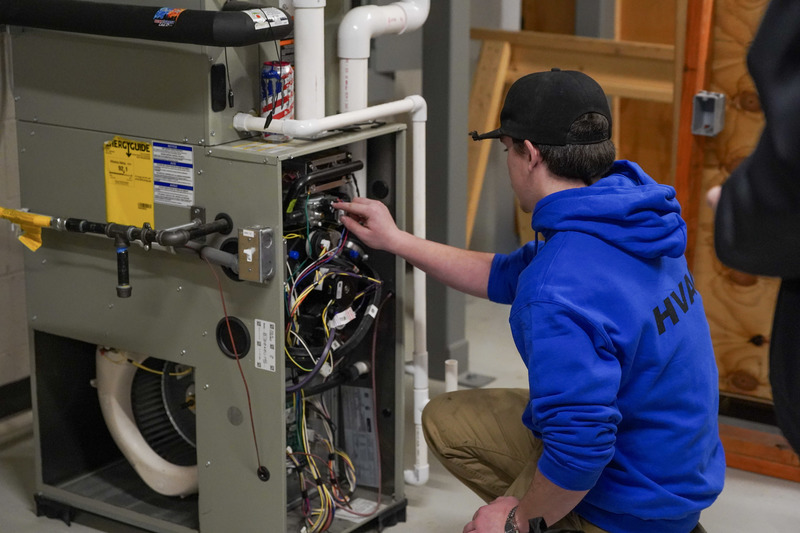 A student in a blue sweatshirt kneels in front of an AC unit observing the wiring.