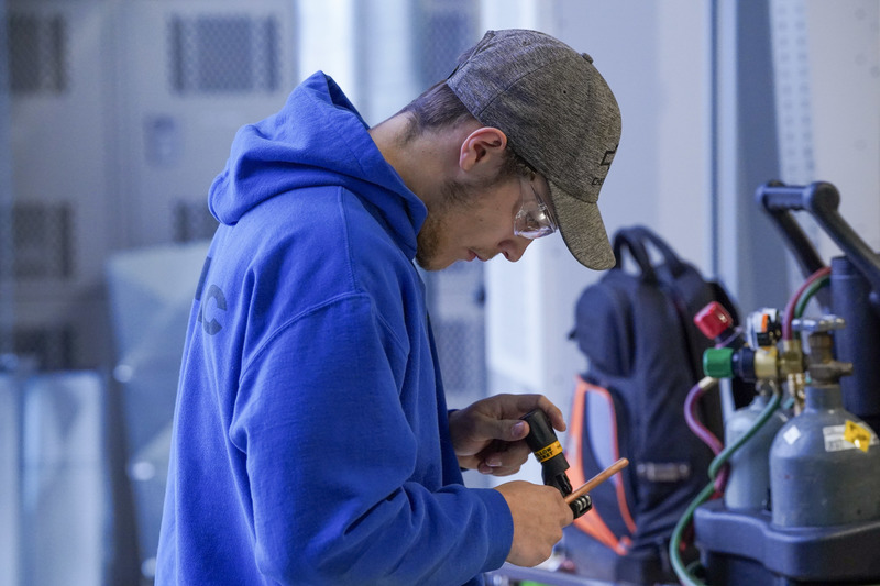 A student in a blue sweatshirt wears work glasses and examines a piece of coil in his hand.