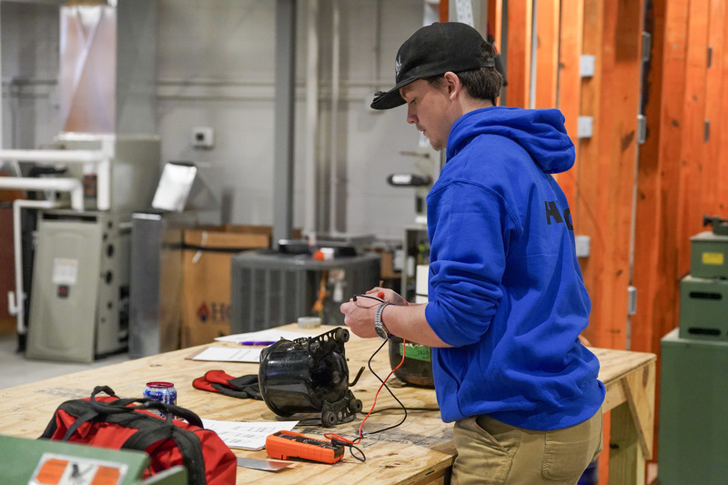 A student in a blue sweatshirt stands at a work bench working on an air compressor.