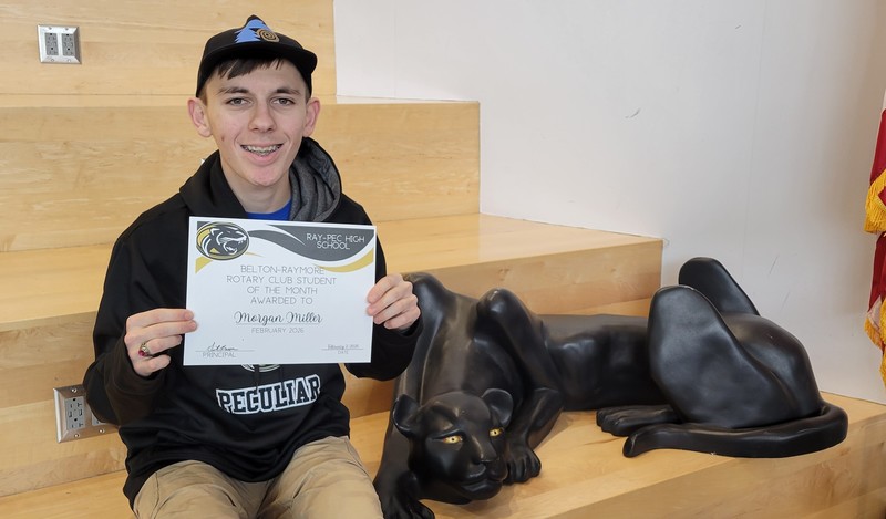 Student wearing a hat and black sweatshirt holding certificate for Student of the month posing with panther on a set of stairs.