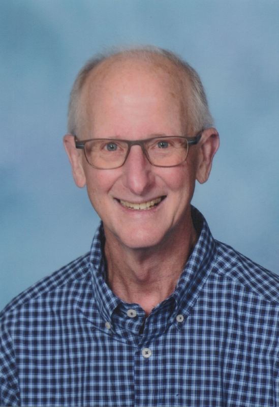 An older gentleman with glass and a plaid shirt stands infront of a blue backdrop smiling
