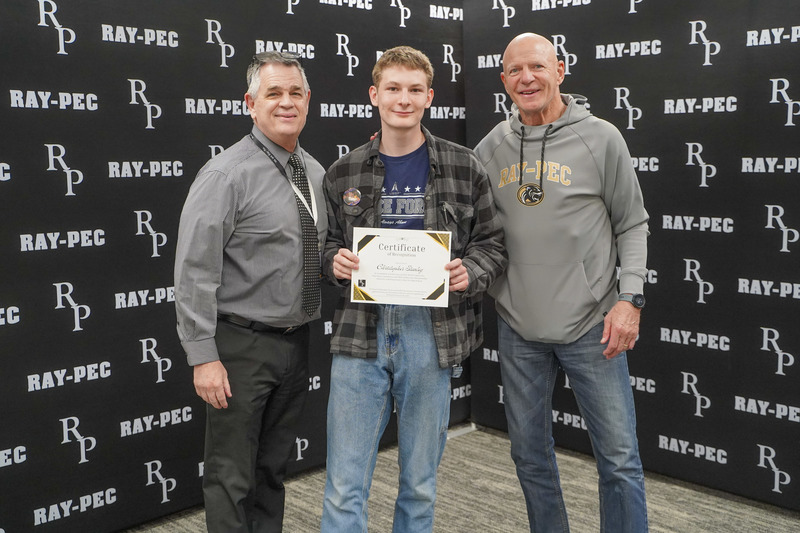 Three men stand together with the boy in the middle holding up a certificate