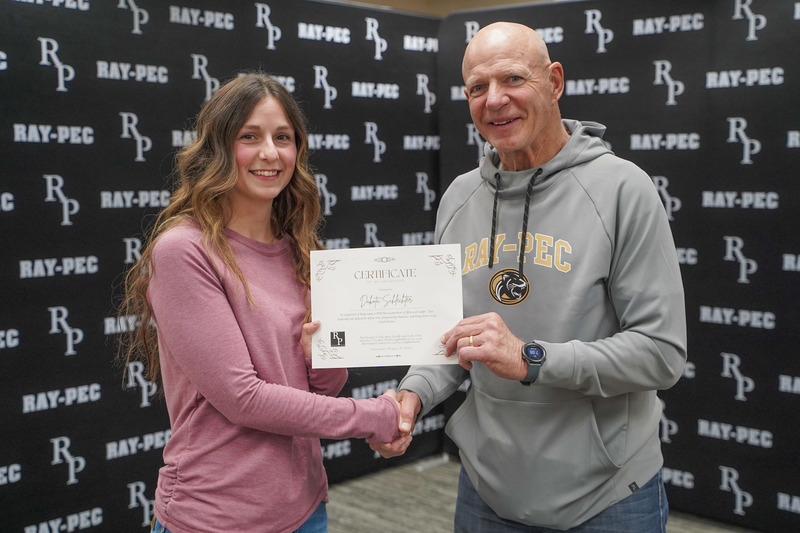 A man and woman stand together shaking hands while holding a certificate