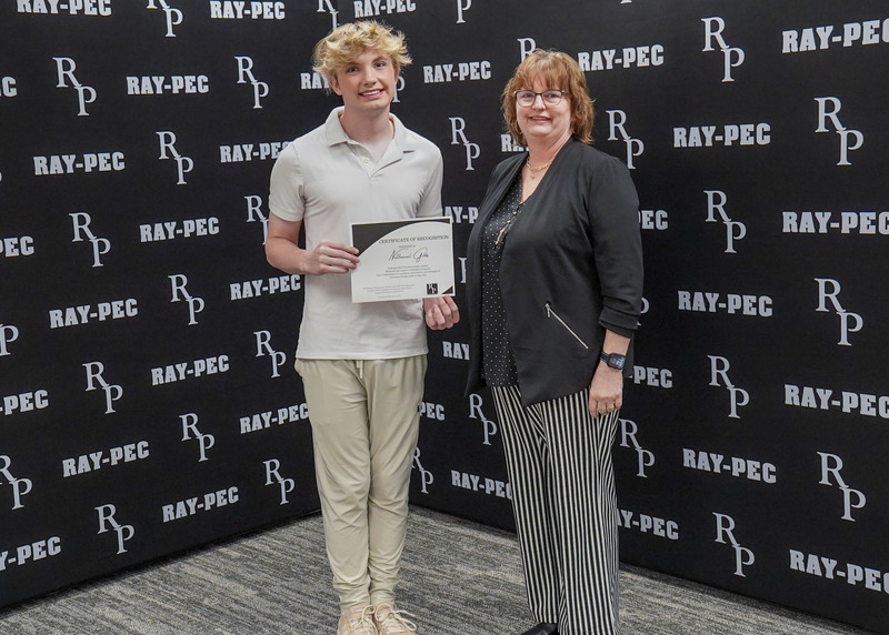 A boy and woman stand in front of a black backdrop with the boy holds a certificate