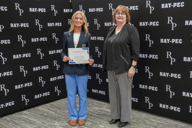 A girl and woman stand in front of a black backdrop with the girl holds a certificate