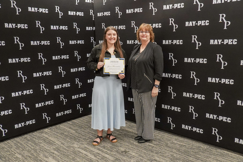 Two women stand in front of a black backdrop with one of the women holding a certificate