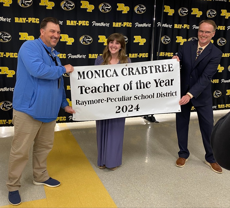 Two men hold a white banner with a woman standing behind it smiling. The banner reads, "Monica Crabtree Teacher of the Year 2024"