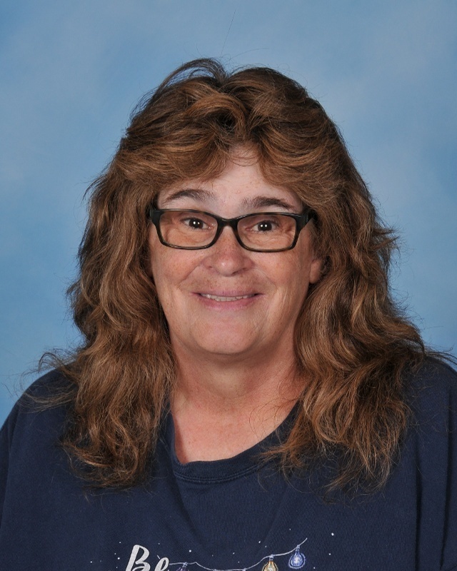 A woman with glasses, her hair down, and a blue shirt smiles in front of a blue backdrop