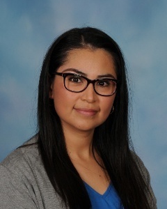 A woman with dark hair and glasses smiles in front of a blue backdrop