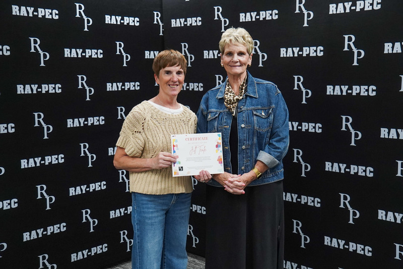 Two woman, one holding a certificate, stand smiling infront of a black backdrop with RP branding.