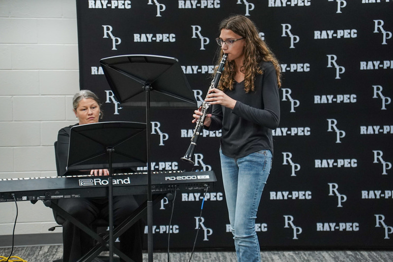 A young musician plays a clarinet with a woman playing the piano behind her.