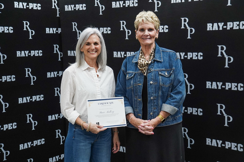 Two woman, one holding a certificate, stand smiling infront of a black backdrop with RP branding.