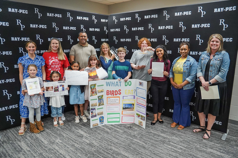 A group of students and teachers all holding projects, stand in front of a black backdrop with RP branding.