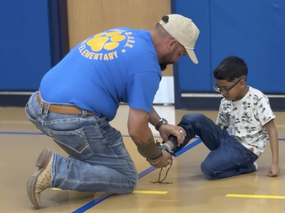 A person wearing a cap, helping a kid tie his shoe, the person has a  blue shirt that says "tiger dad"