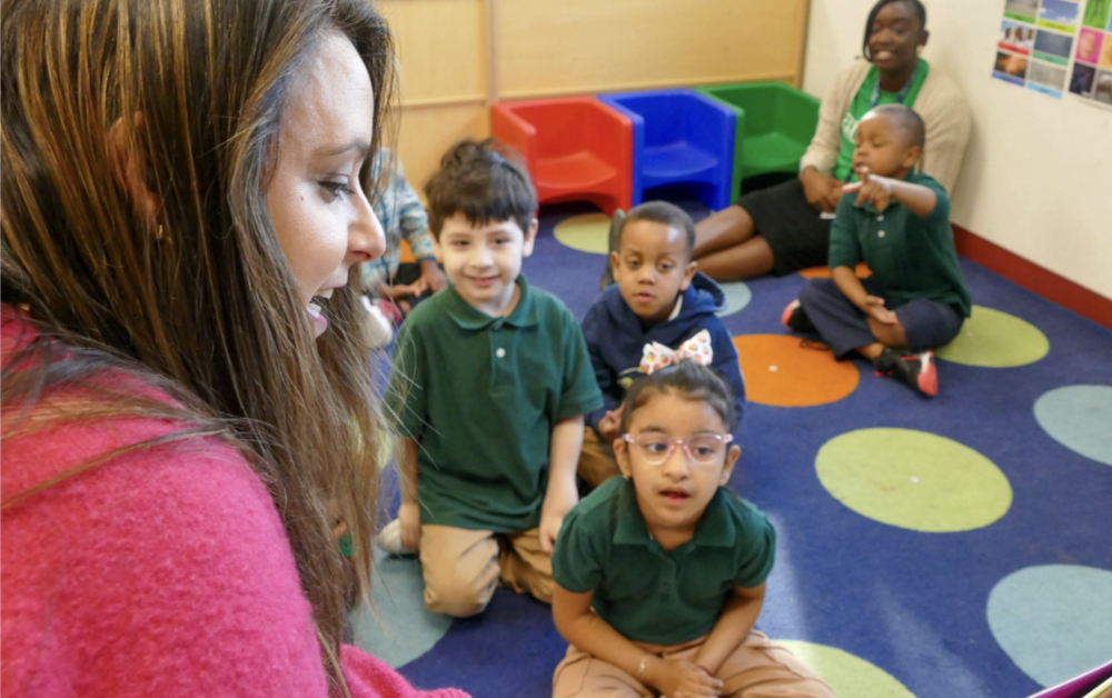 Dr. April Estrada, left side of screen image, sits in a pink sweater in front of a classroom. Dr. Estrada reads a lesson to a group of very young students who all look eager to learn, while sitting on a blue rug with dots. 