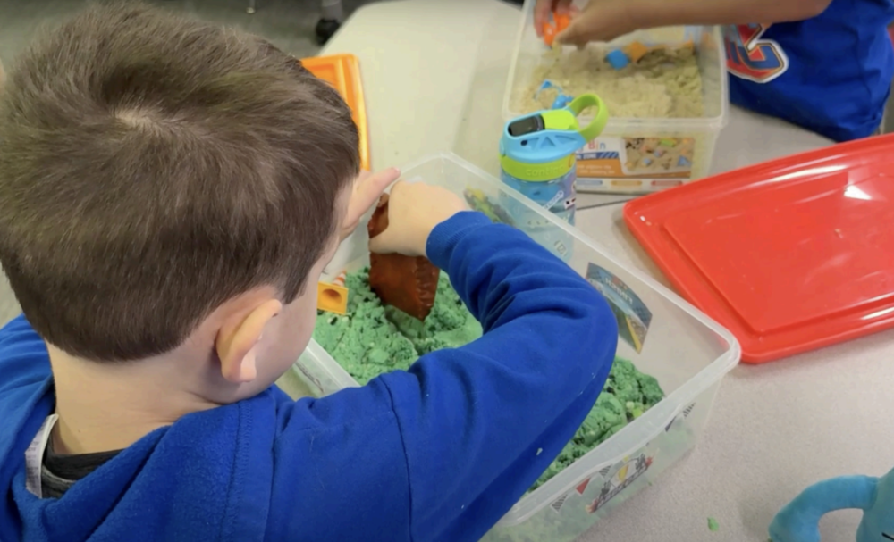 Young boy, student, playing with green putty or playdough in his classroom.