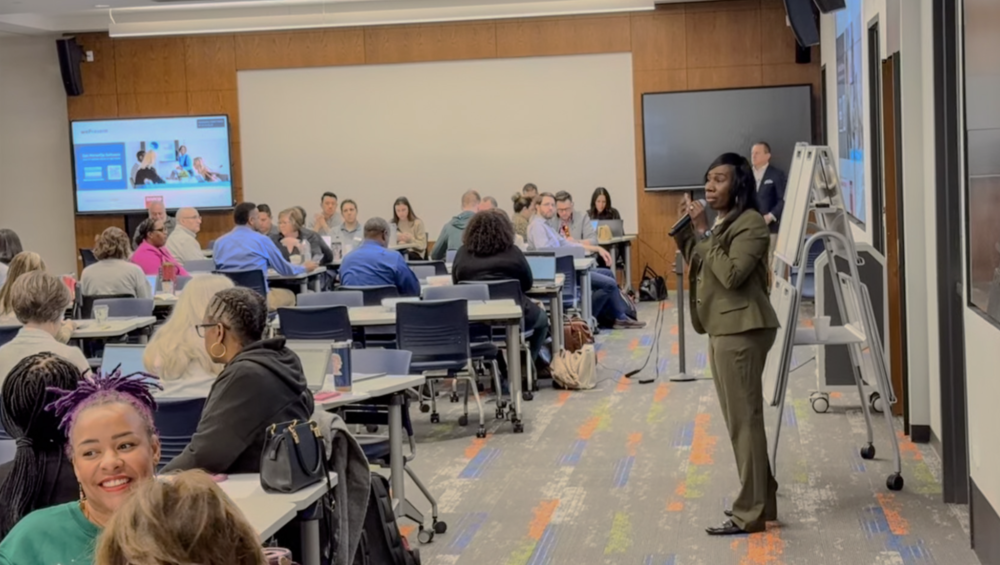 Woman speaking in front of a professional group of individuals in a conference room style set up. All people in the room are engaged in conversation. 
