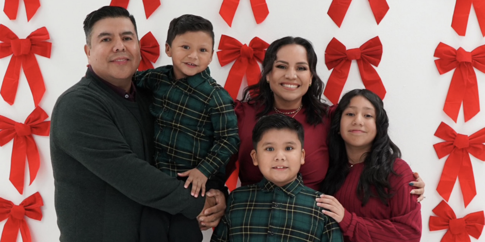 Hispanic family standing in front of a white wall with red holiday bows, the family is in their best holiday attire.