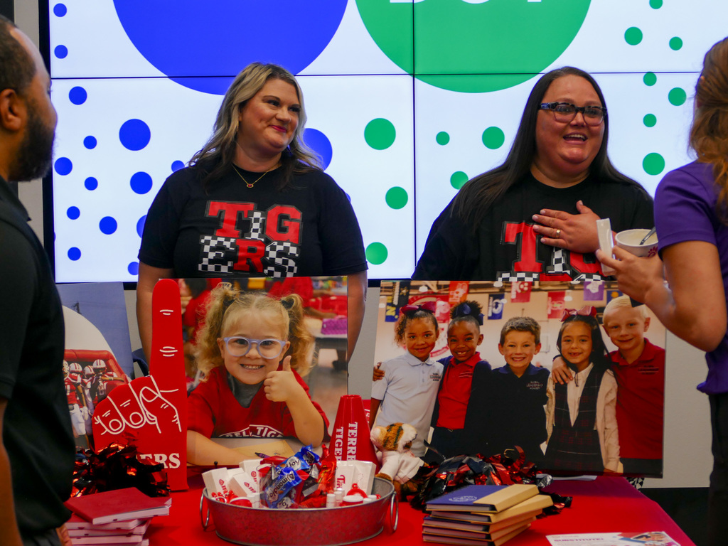 Two individuals smiling behind a promotional table decorated with red items and a photograph featuring a group of children. The setting includes a backdrop with a polka dot design.