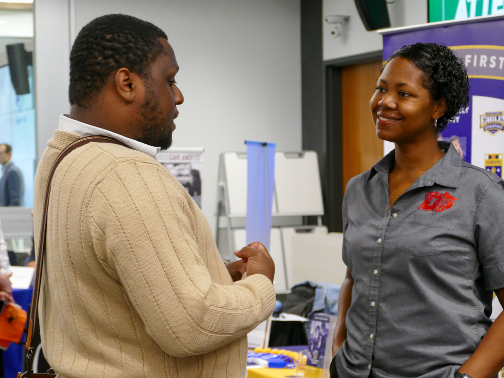 Two individuals are engaged in a conversation at an event, with banners and promotional materials visible in the background. One person is wearing a sweater and the other a shirt with a Red Cross logo.